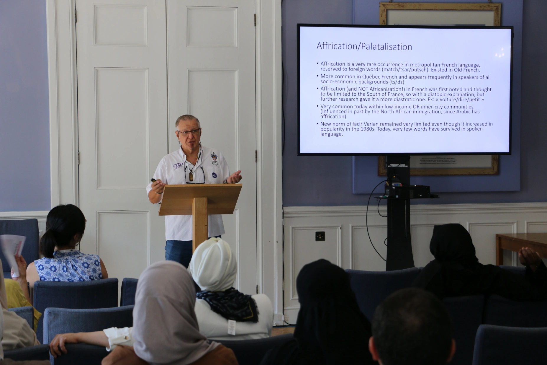 A presenter speaking at a lectern beside a screen showing slides, with an audience seated in front
