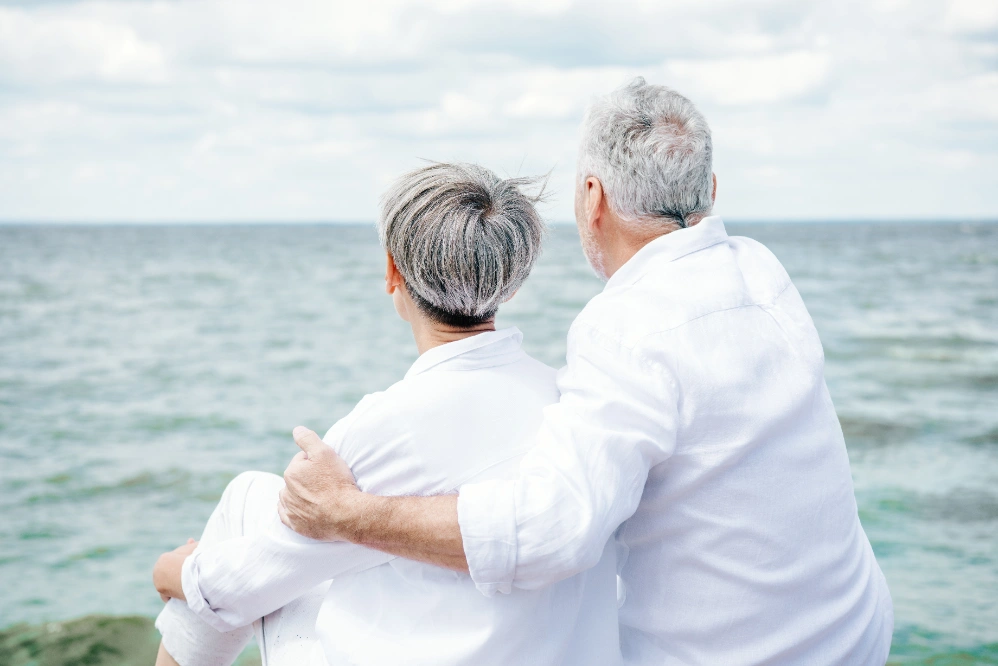 An older couple in white shirts sitting by the sea, his arm around her shoulders