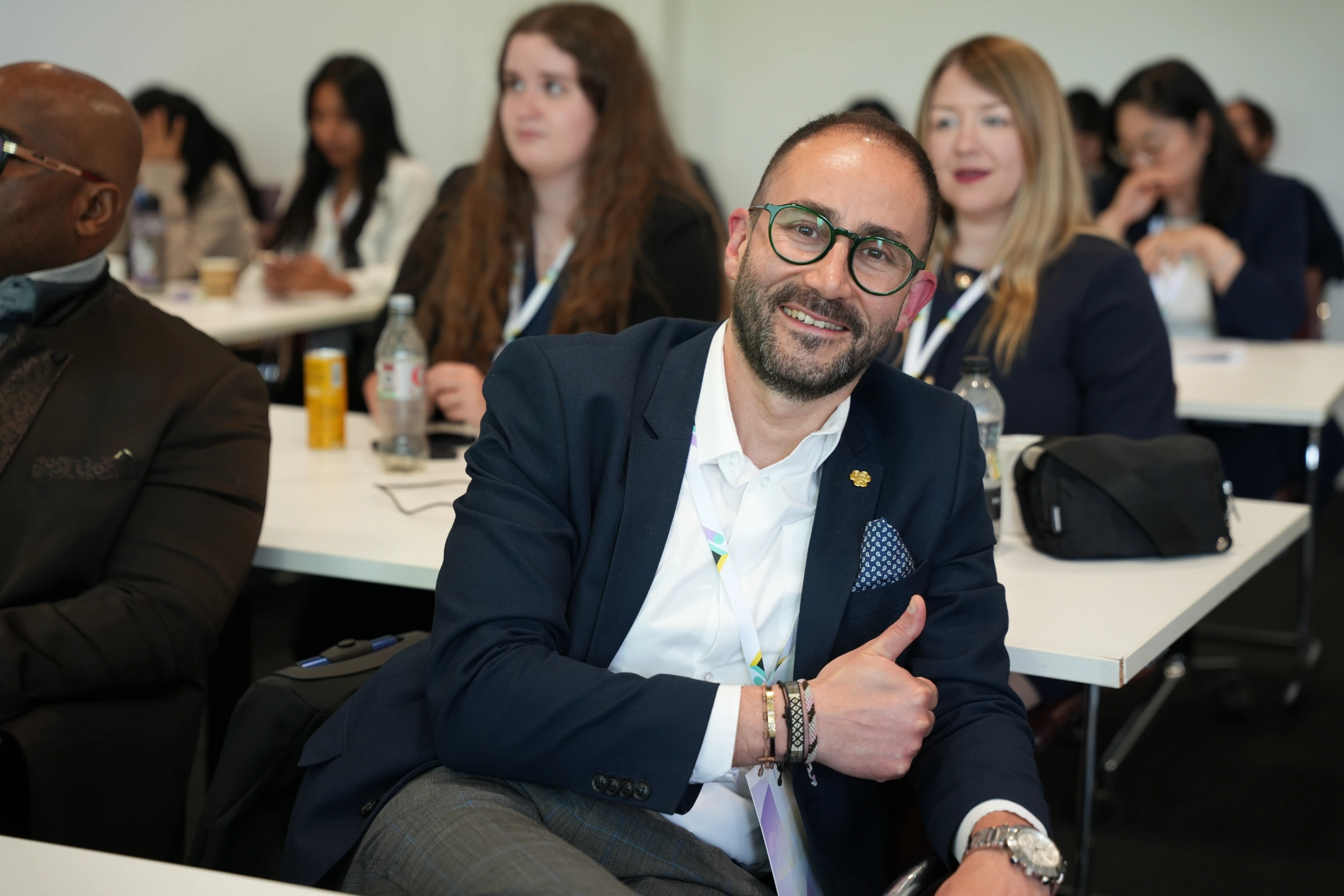 A smiling conference participant giving a thumbs up, seated among other attendees